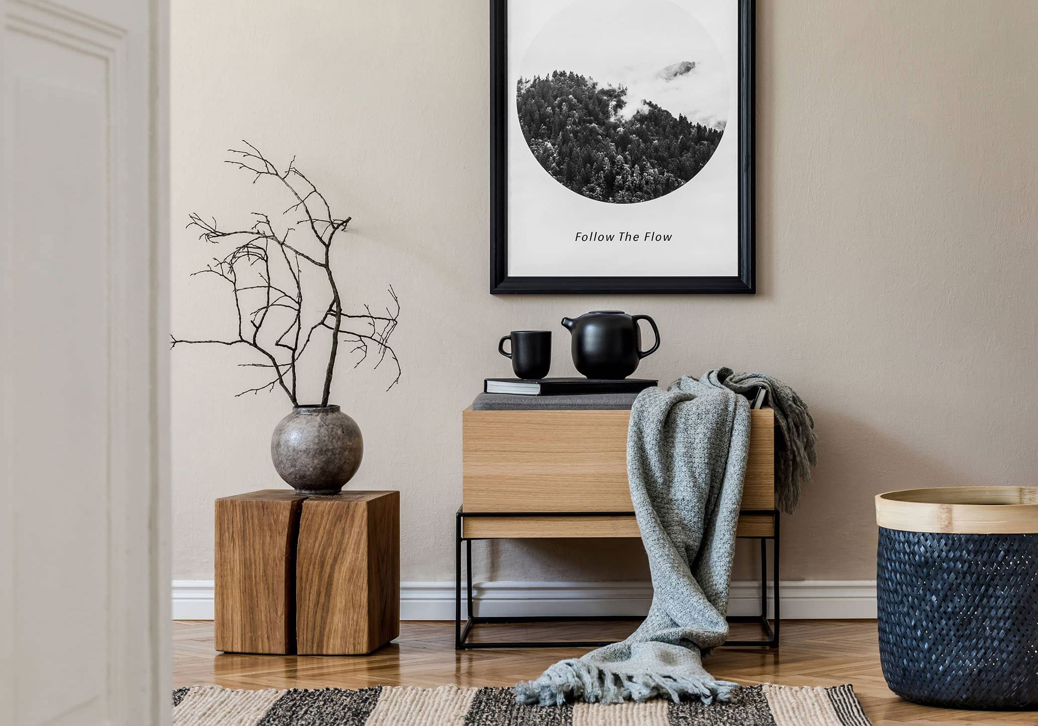 Architecturally pleasing home decor against a wall in a home including a small table with a soft blanket draping off the side, a frail plant with no leaves and a warm painting that says "Follow The Flow"